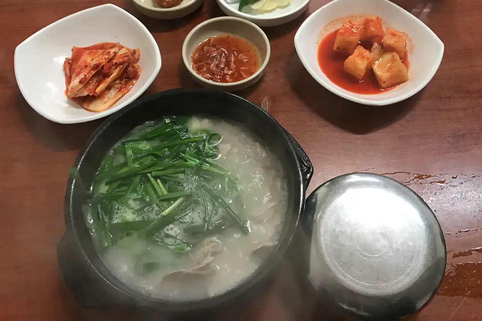 A stone bowl of dwaeji gukbap topped with chives, with kimchi, radish kimchi and a small dish of salted shrimp. Similar to a Busan bowl, not from a shop named on this page.