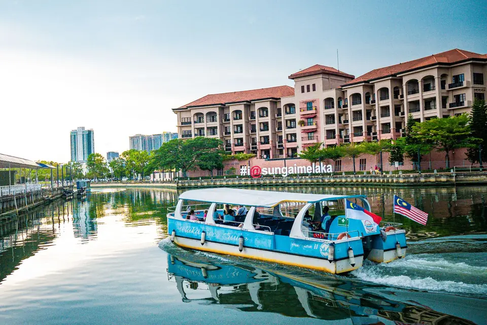 A river cruise boat on the Melaka River, passing red-roofed buildings on a calm afternoon