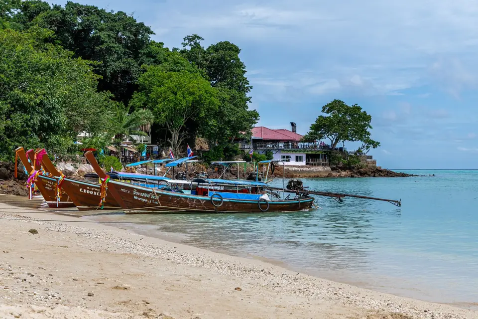 Wooden longtail boats pulled up on a quiet beach in southern Thailand, with turquoise water beyond