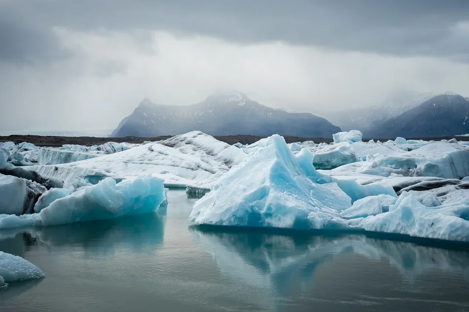 Blue-white icebergs floating in still glacier lagoon water, with snowy mountains under low cloud behind