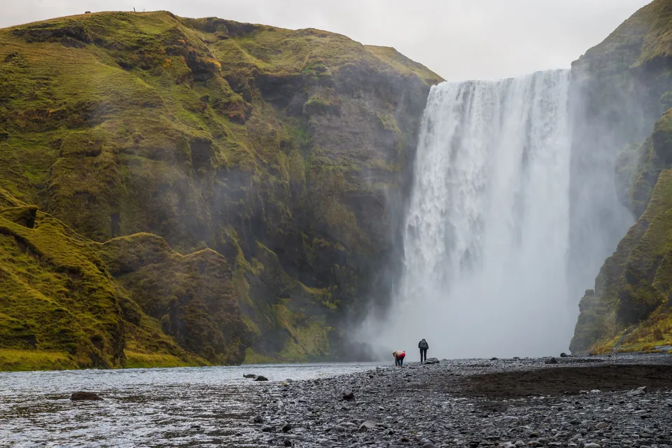 Two visitors on the stony riverbank below Skógafoss waterfall in south Iceland, between mossy green cliffs