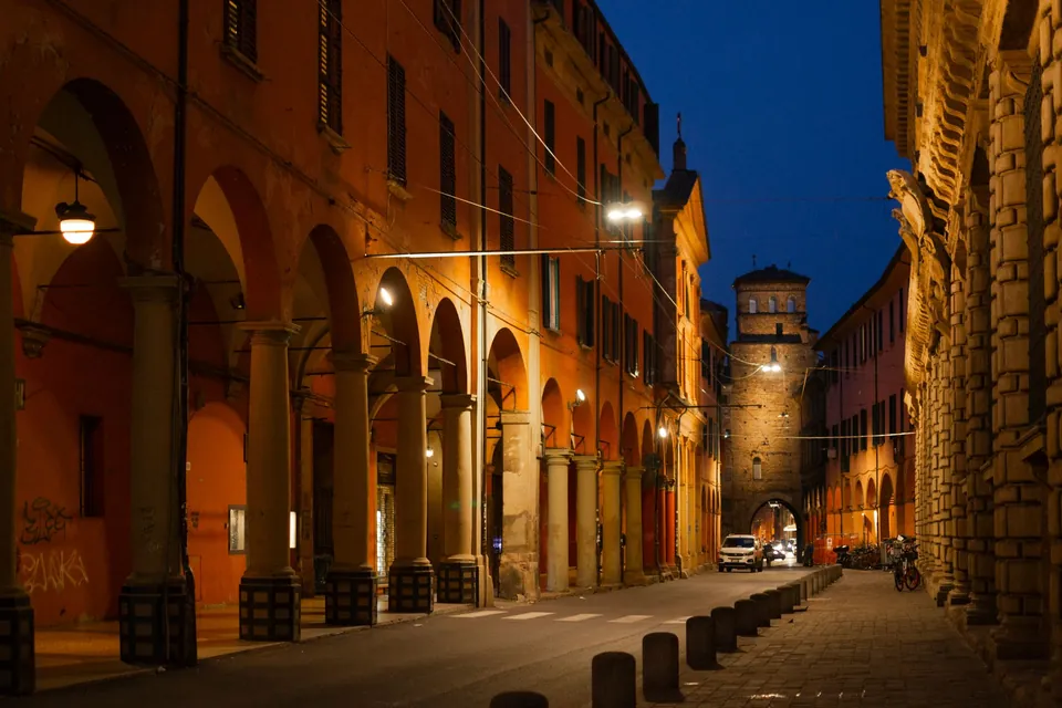 A lit portico in Bologna in the evening, with an old brick tower at the end of the street