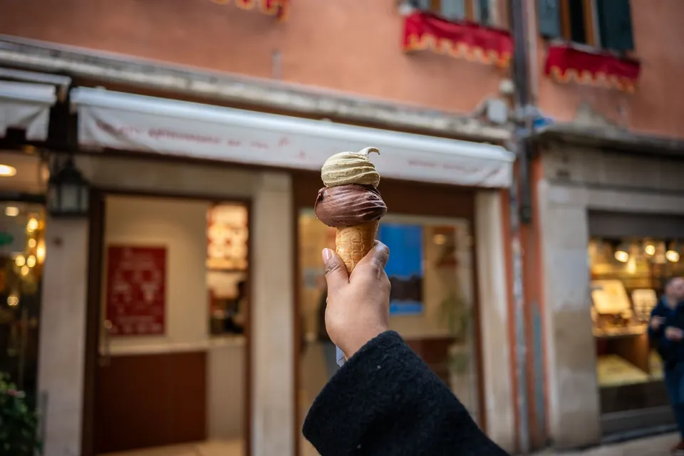 A hand holding a gelato cone on a blurred street in Venice
