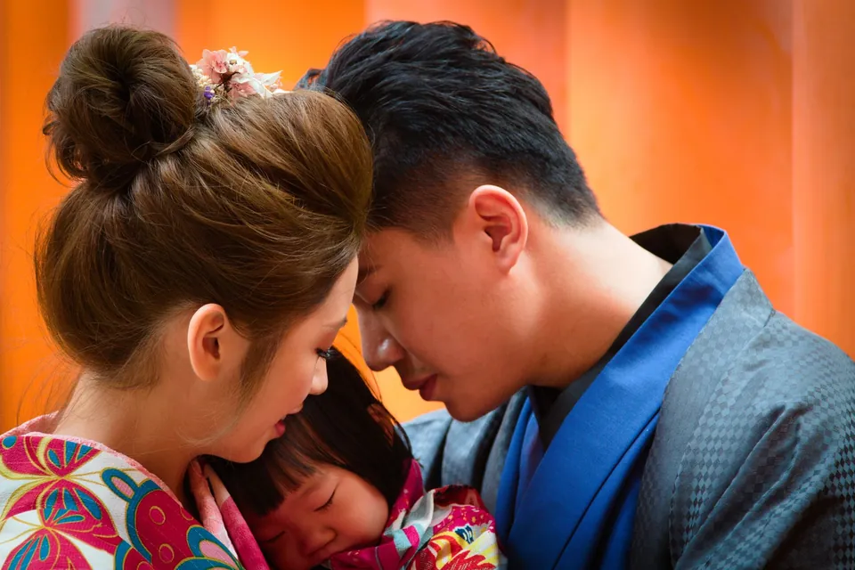 Parents and a small child in colourful kimonos sharing a quiet hug against an orange background