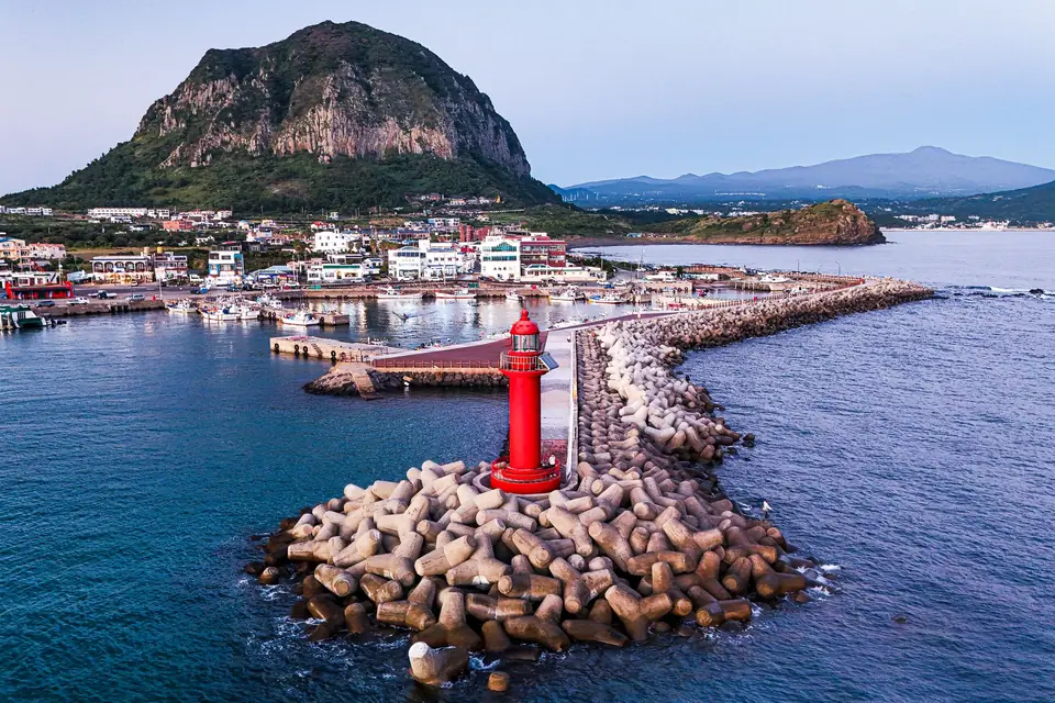A red lighthouse on a harbour breakwater below Sanbangsan on Jeju's southwest coast, with Hallasan in the distance